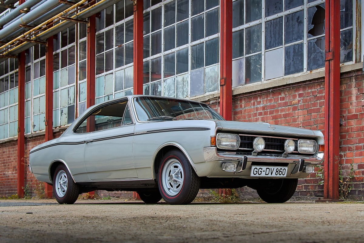 Opel Kadett B versão Rallye com faixas desportivas, fotografado em ambiente de competição histórica, com público ao fundo